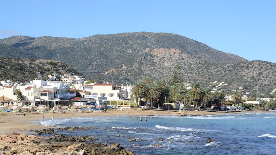 A coastal town with buildings, a beach, rocky shoreline, and a mountain in the background.