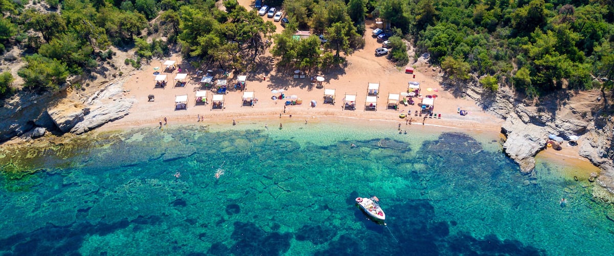 An aerial view of a beach with clear turquoise waters, a small boat anchored near the shore, and several beach huts and tents.
