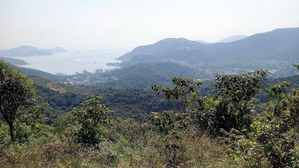 A mountainous landscape with dense greenery, a distant body of water, and a small settlement.