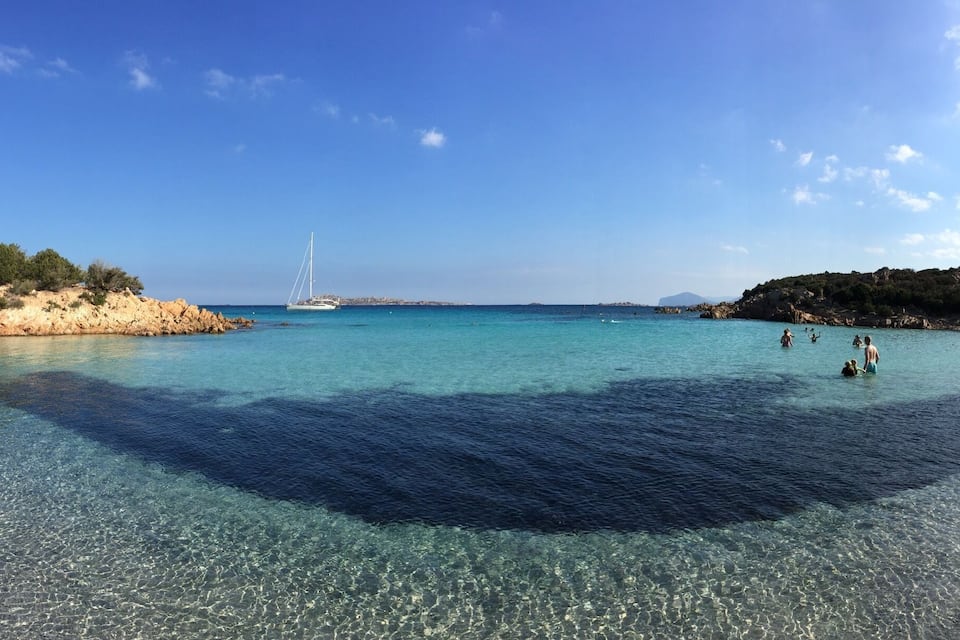 A clear coastal beach with rocky shores, a sailboat in the distance, and people swimming.
