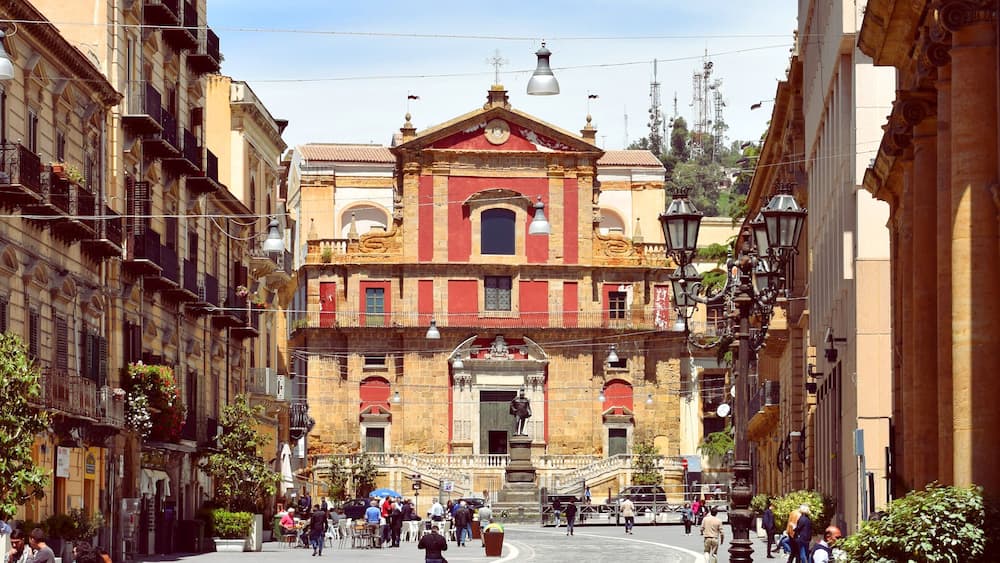 Une place historique, dominée par une église remarquable à la façade rouge et or, et entourée d’édifices représentatifs de l’architecture européenne classique.