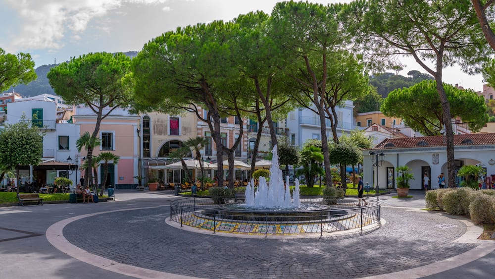 A cobblestone plaza with a central fountain, surrounded by trees and buildings with Mediterranean architecture.