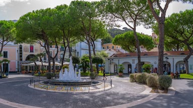 Una piazza lastricata con una fontana al centro, circondata da alberi e edifici dall’architettura mediterranea.