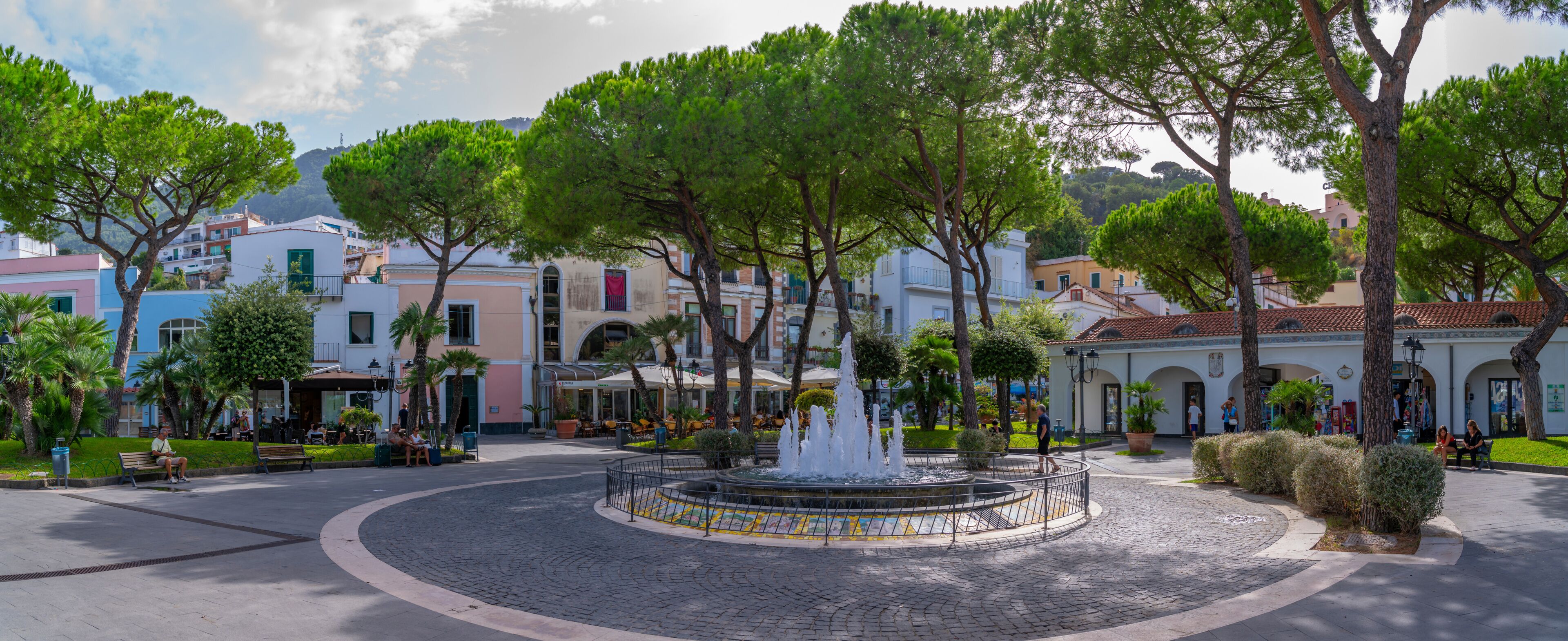 A cobblestone plaza with a central fountain, surrounded by trees and buildings with Mediterranean architecture.