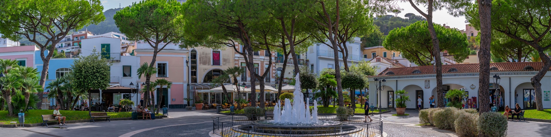 A cobblestone plaza with a central fountain, surrounded by trees and buildings with Mediterranean architecture.