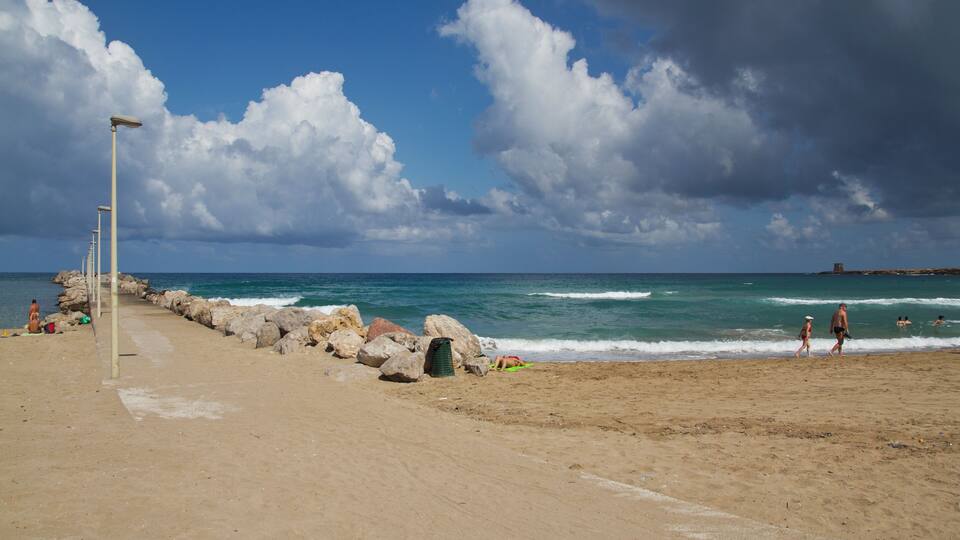 A sandy beach with a rocky breakwater, calm sea, and a clear sky with clouds.