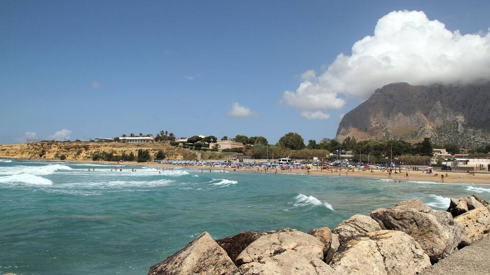A beach with waves, umbrellas, and a mountain in the background.