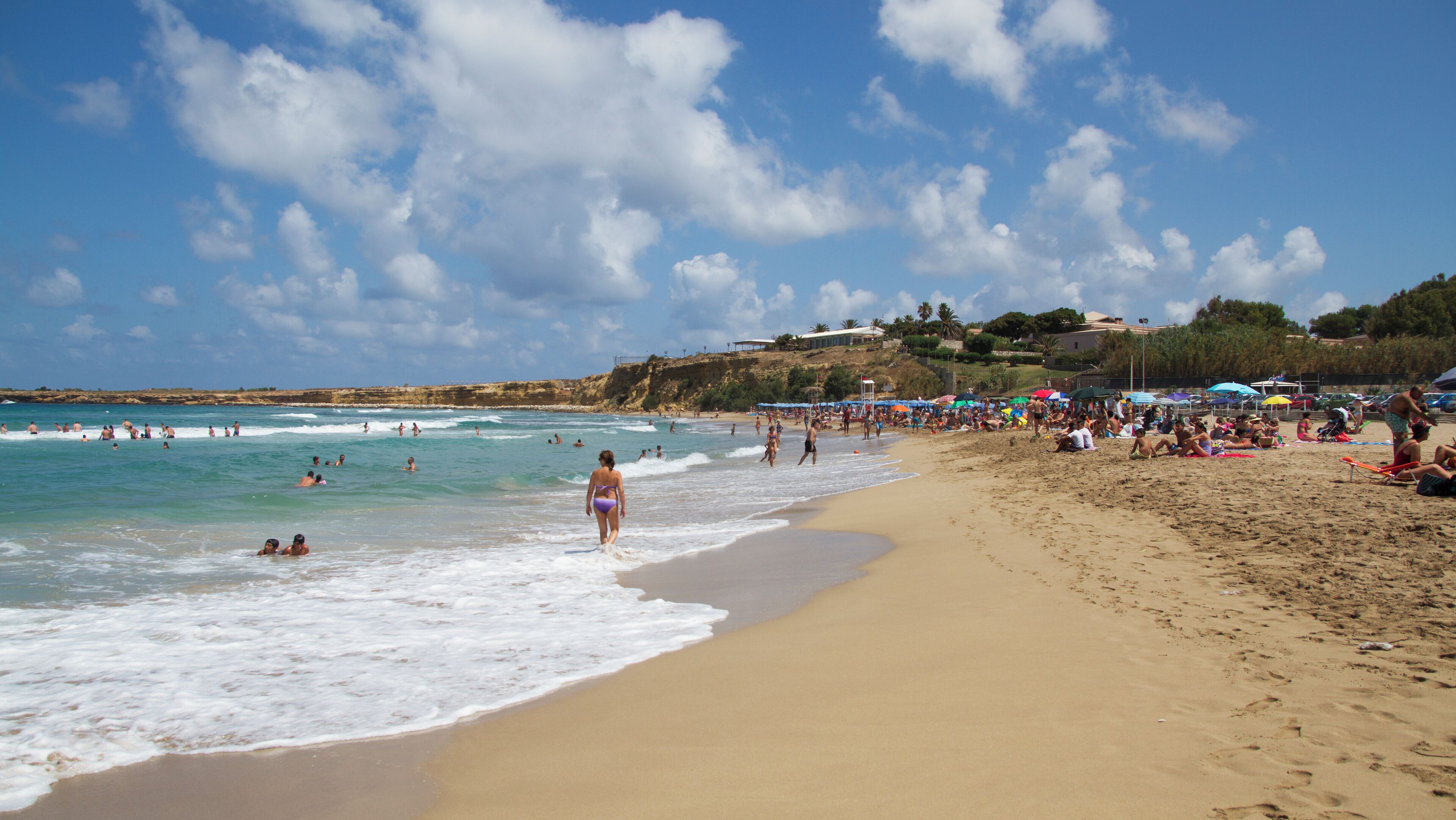 A crowded beach with people swimming and sunbathing, a sandy shore, and a clear blue sky with clouds.
