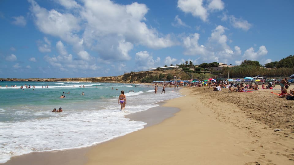 A crowded beach with people swimming and sunbathing, a sandy shore, and a clear blue sky with clouds.