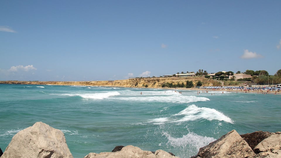 A coastal beach with waves, rocky outcrops, and a building on a cliff.