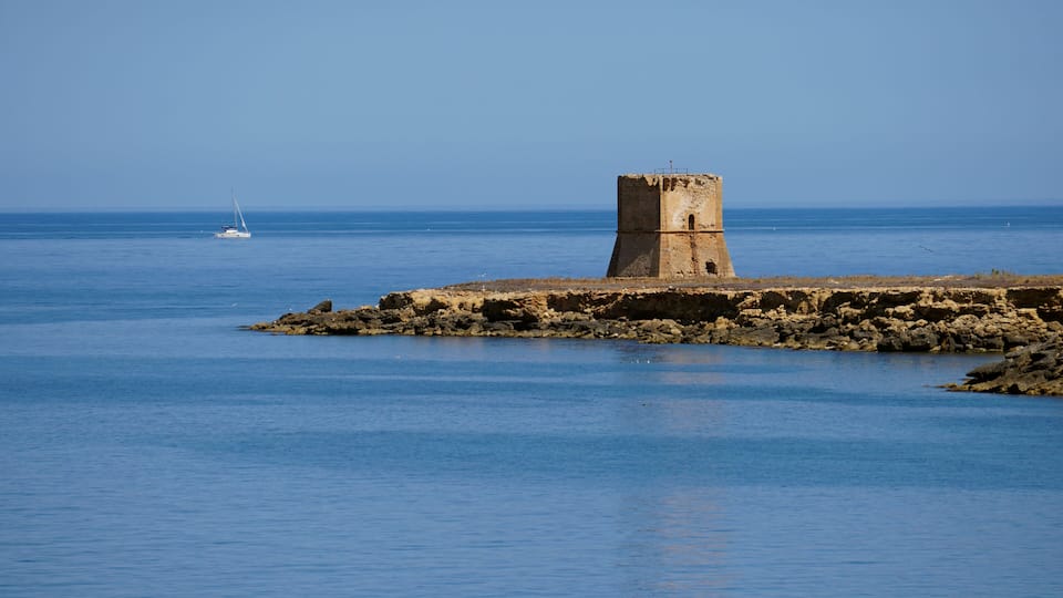 A coastal tower on a rocky outcrop with calm blue waters and a sailboat in the distance.
