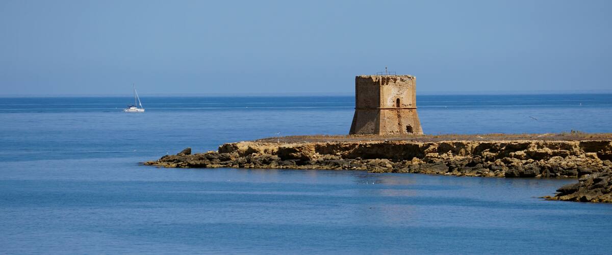 A coastal tower on a rocky outcrop with calm blue waters and a sailboat in the distance.