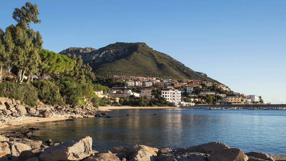 A coastal town with buildings on a hillside, a rocky shoreline, and calm sea.