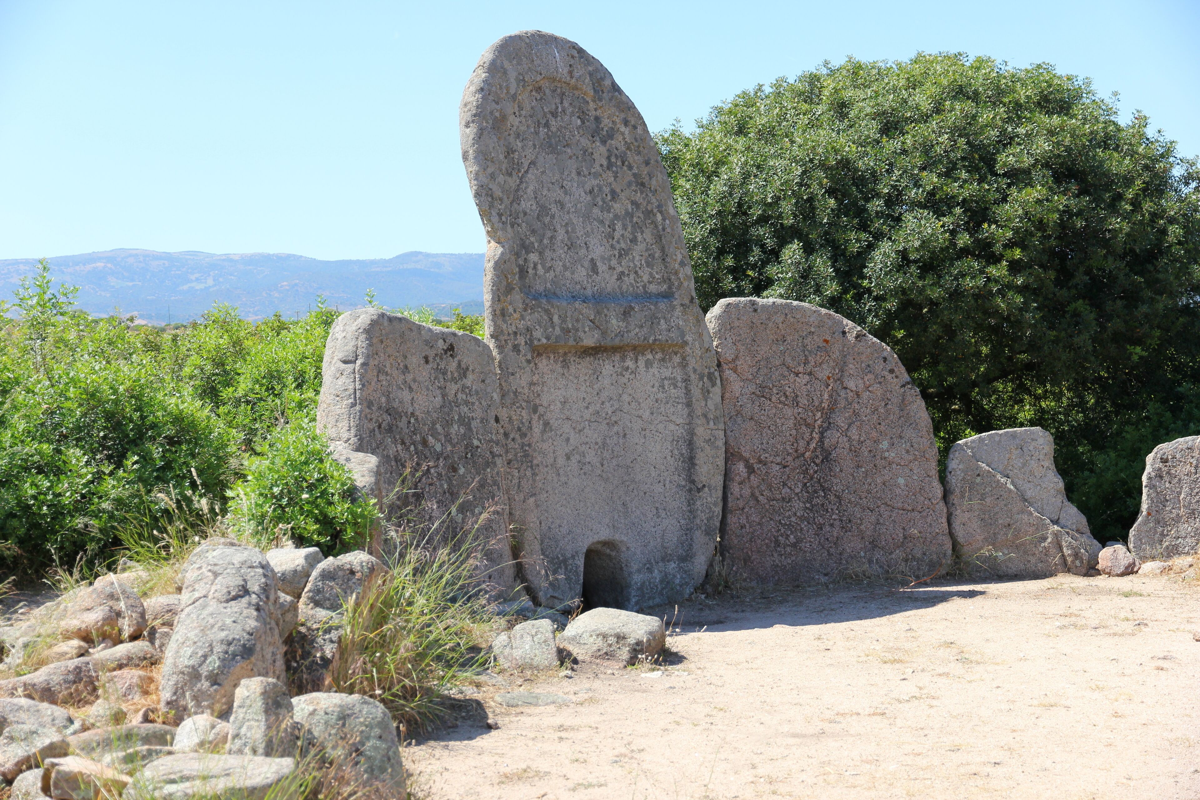 A large stone structure with a carved figure, surrounded by rocks and greenery.