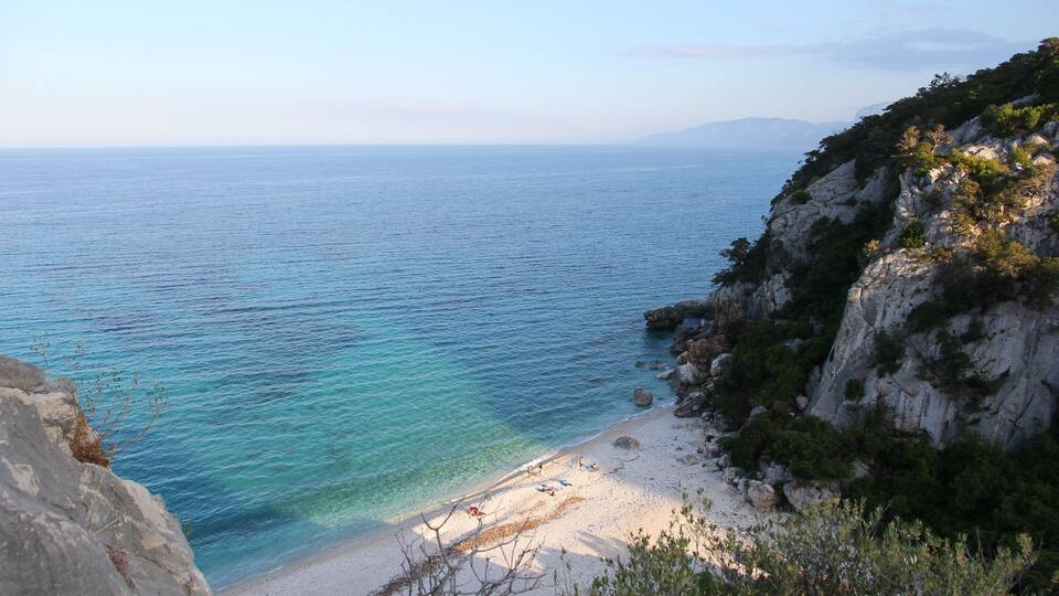 A coastal landscape with a clear blue sea, a sandy beach, and rocky cliffs.