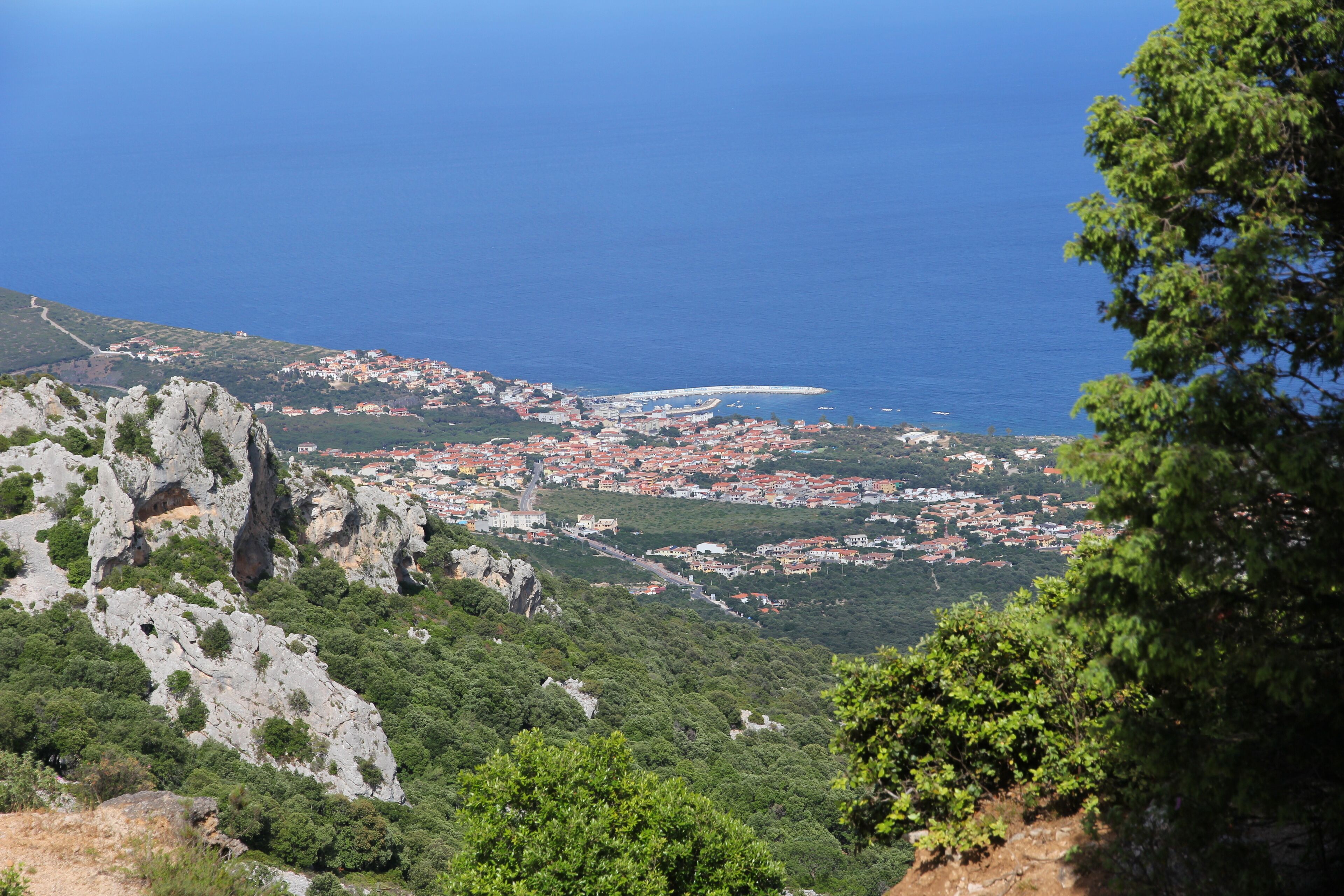 A coastal town with red-roofed buildings, a rocky hillside, and a clear blue sea.