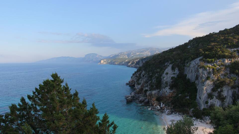 A coastal landscape with clear turquoise waters, rocky cliffs, and dense greenery.