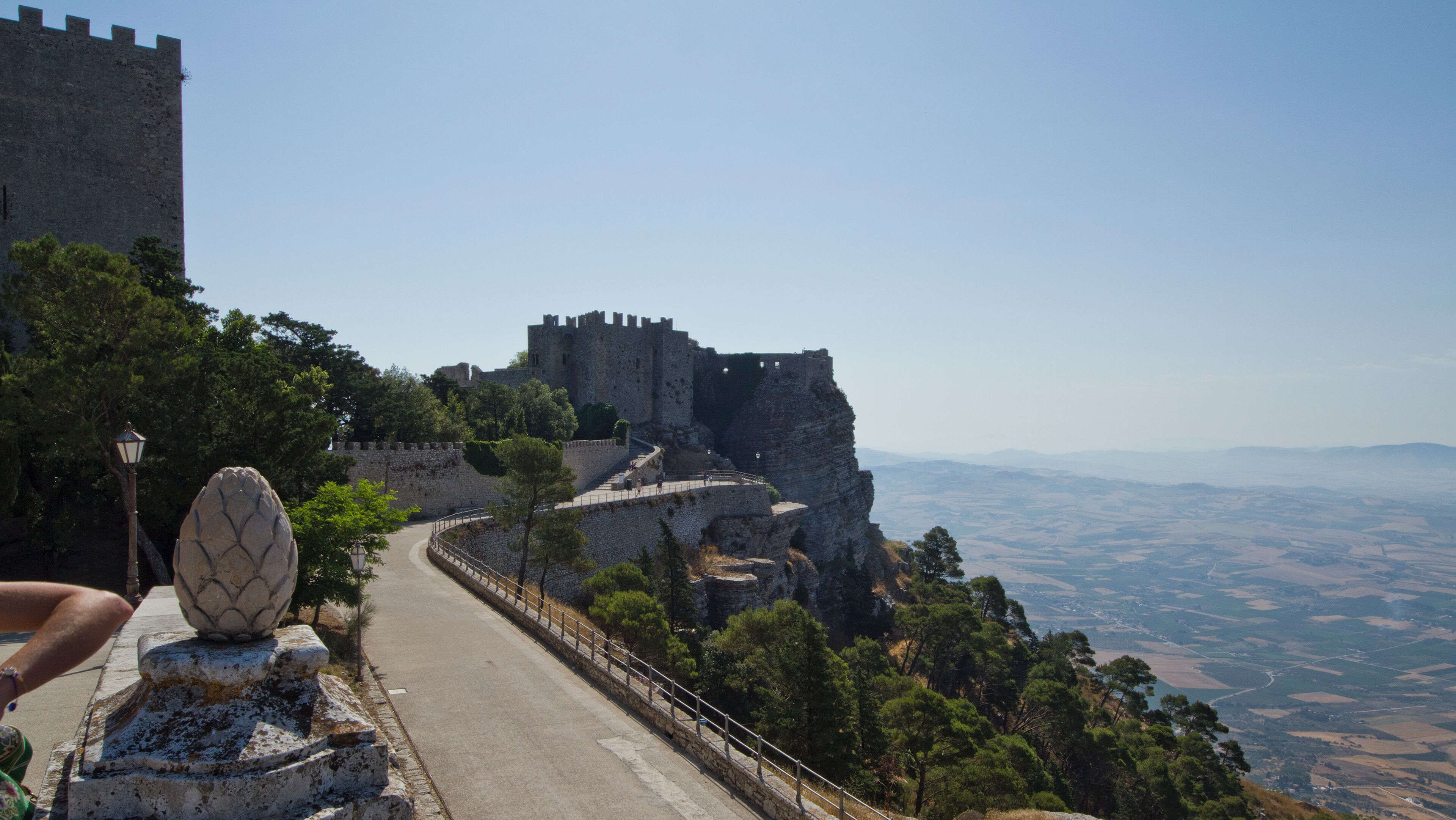 A stone pathway leading to a historic castle atop a cliff, overlooking a valley.