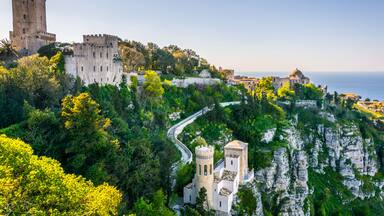 A historic castle on a cliff overlooking the sea, surrounded by lush greenery and a winding road.