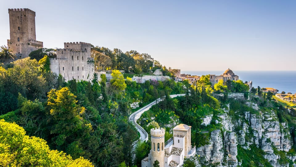 A historic castle on a cliff overlooking the sea, surrounded by lush greenery and a winding road.