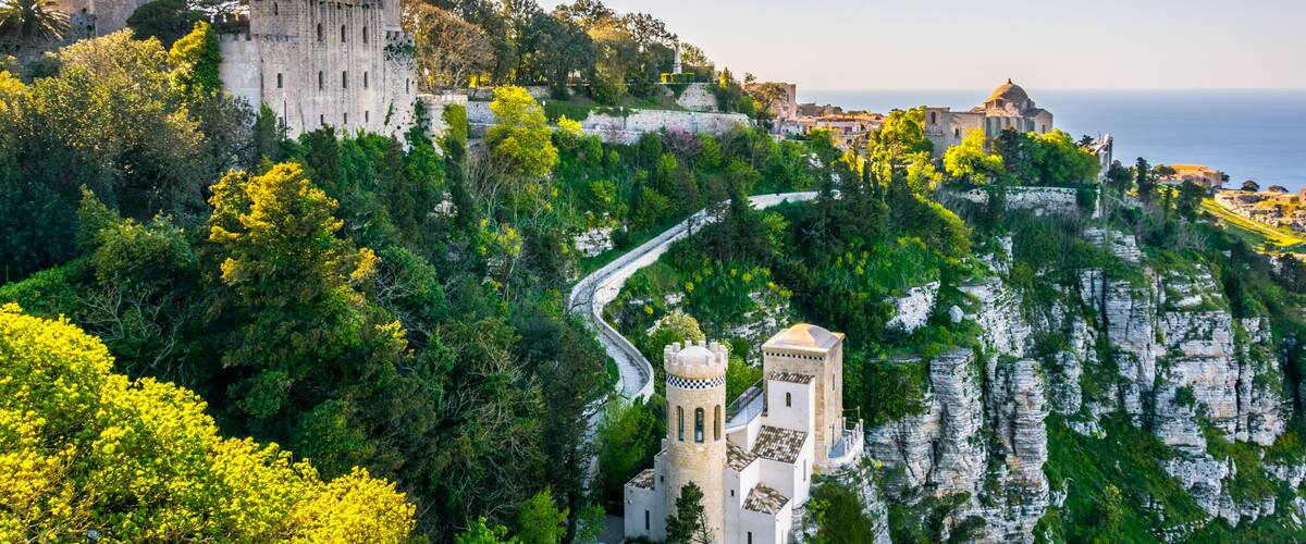 A historic castle on a cliff overlooking the sea, surrounded by lush greenery and a winding road.