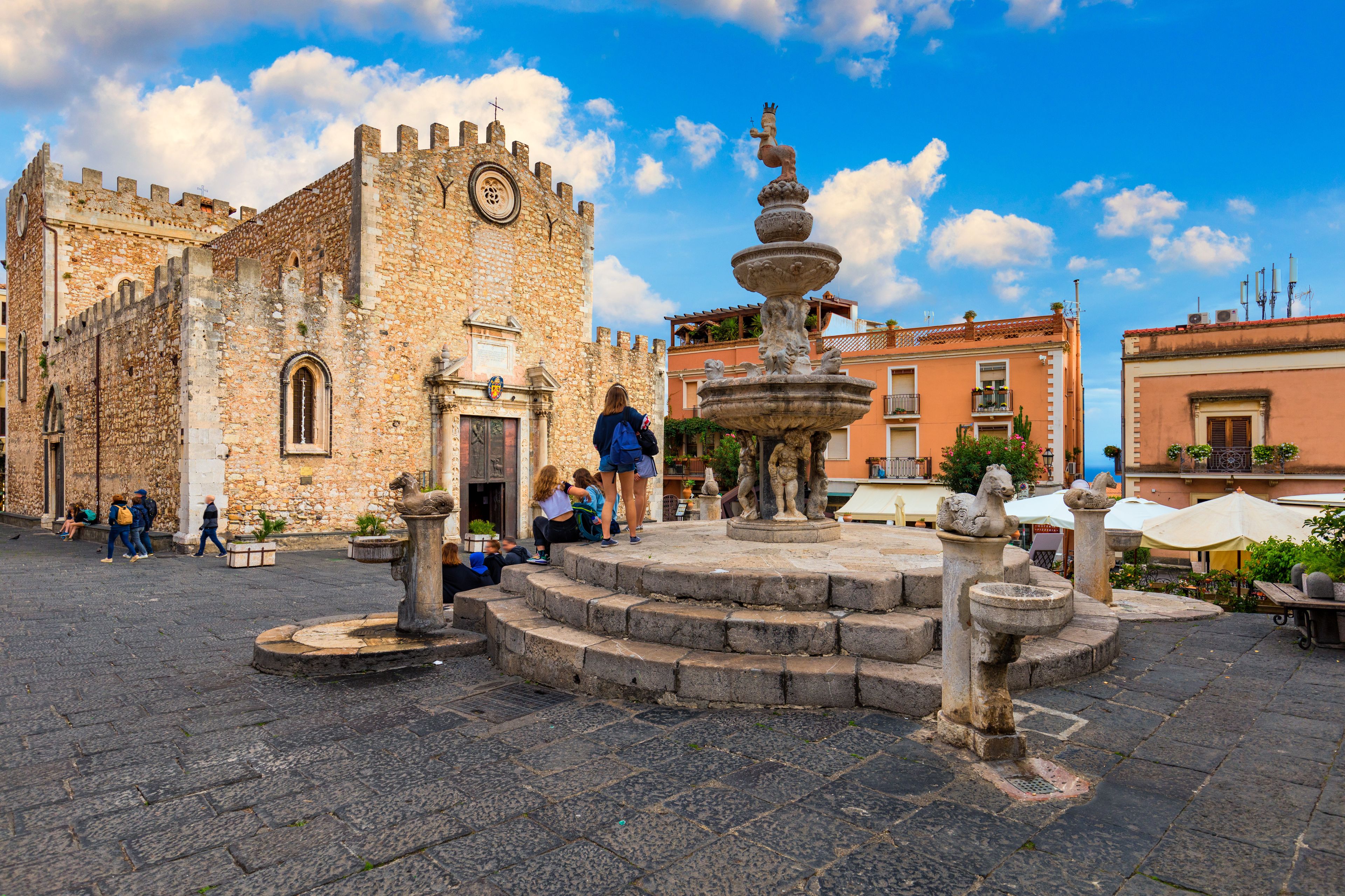 A historic stone building with a tower, a fountain with statues, and a clear blue sky.