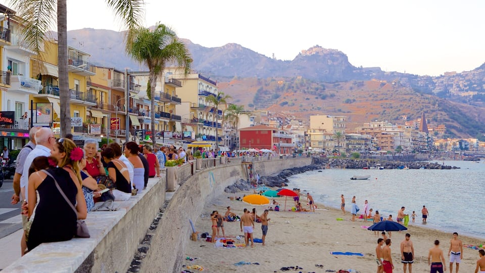 A crowded beach with people sunbathing and swimming, a rocky shoreline, and colorful buildings along the coast.