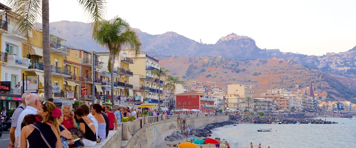 A crowded beach with people sunbathing and swimming, a rocky shoreline, and colorful buildings along the coast.