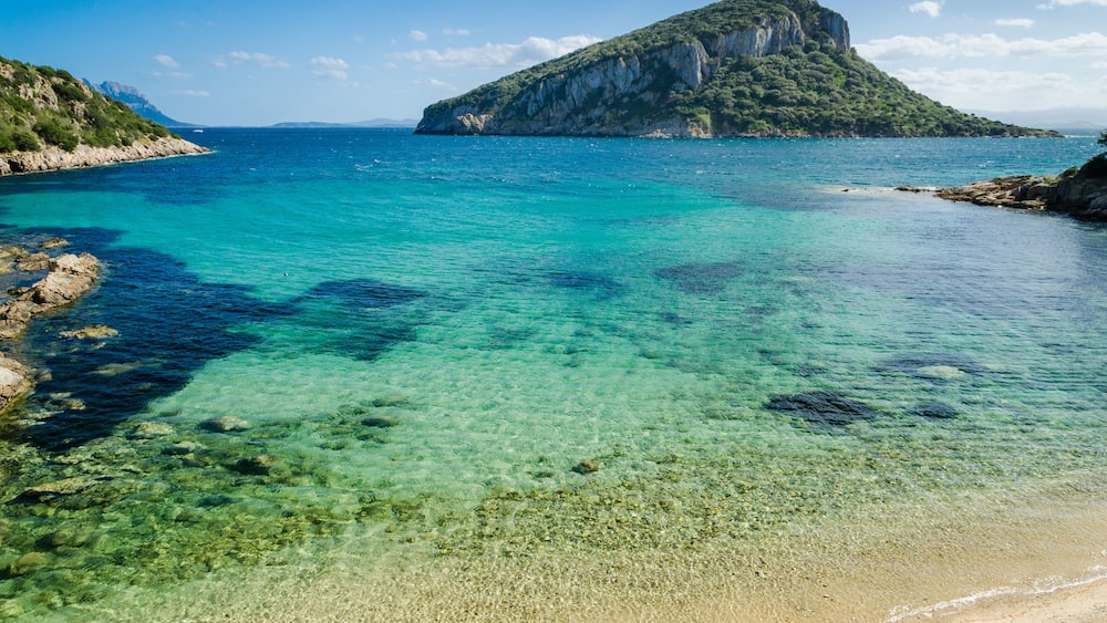 A clear coastal area with turquoise waters, a sandy beach, and a prominent rocky island with a peak.
