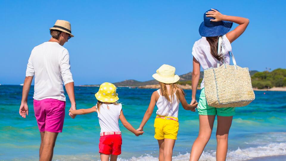 A family of four holding hands on a beach with clear blue water and distant hills.