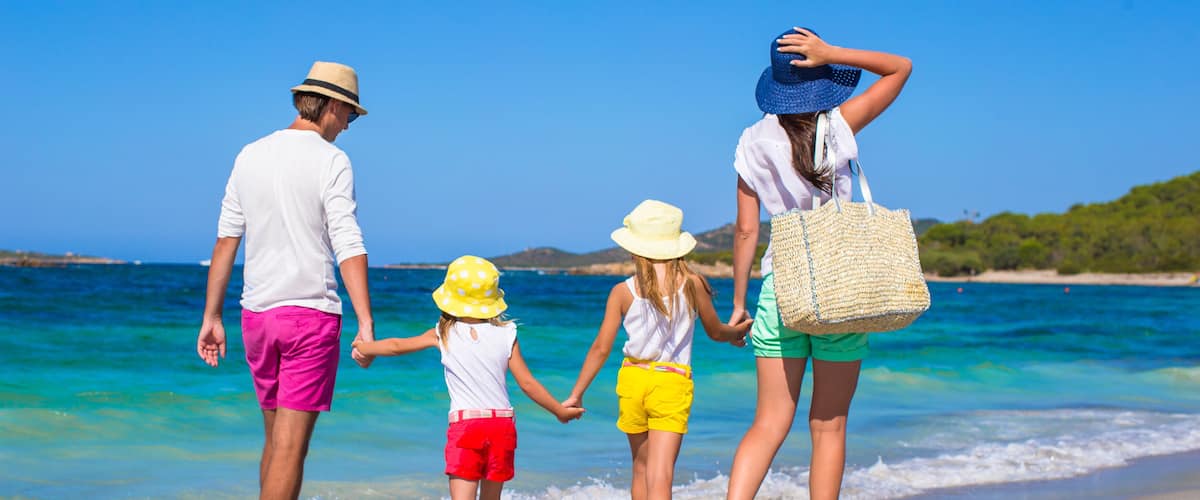 A family of four holding hands on a beach with clear blue water and distant hills.