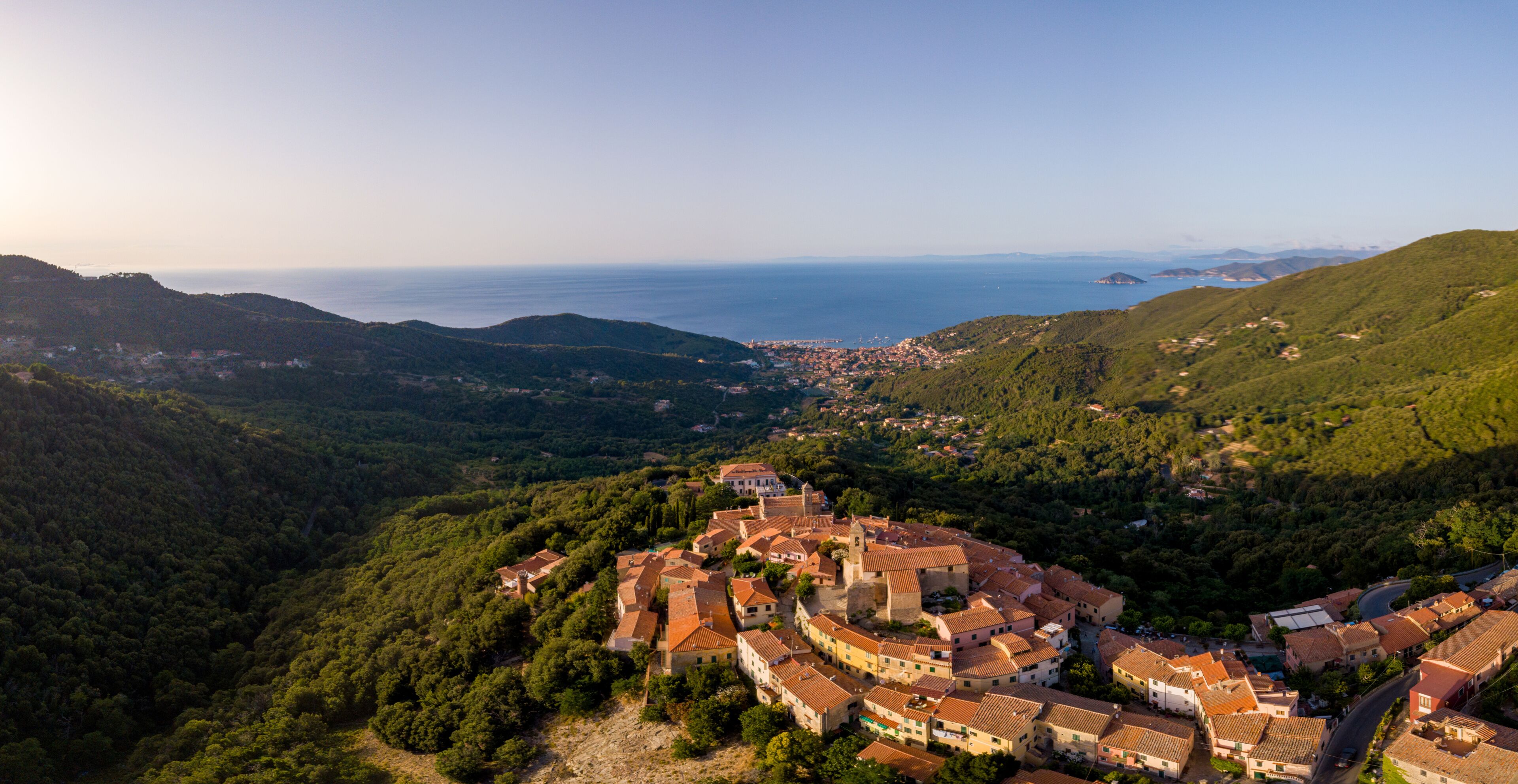 A coastal town nestled in a valley with a prominent castle-like structure surrounded by lush greenery and a view of the sea.