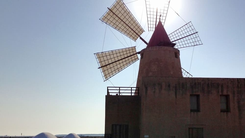 A windmill with a red top and a white sail, located near a body of water with boats.
