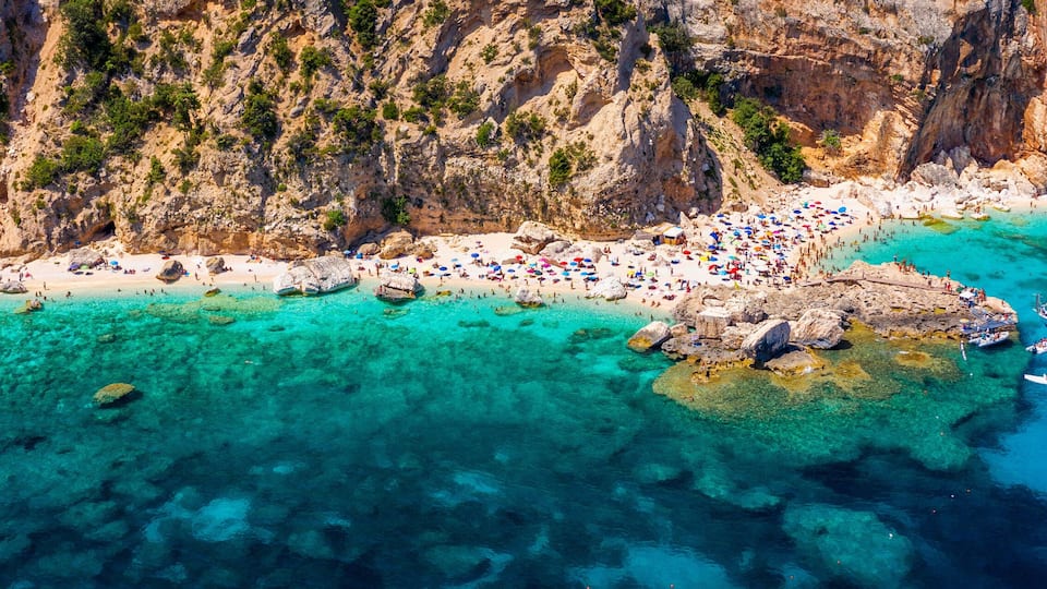 A coastal area with clear turquoise waters, rocky cliffs, and a crowded beach.