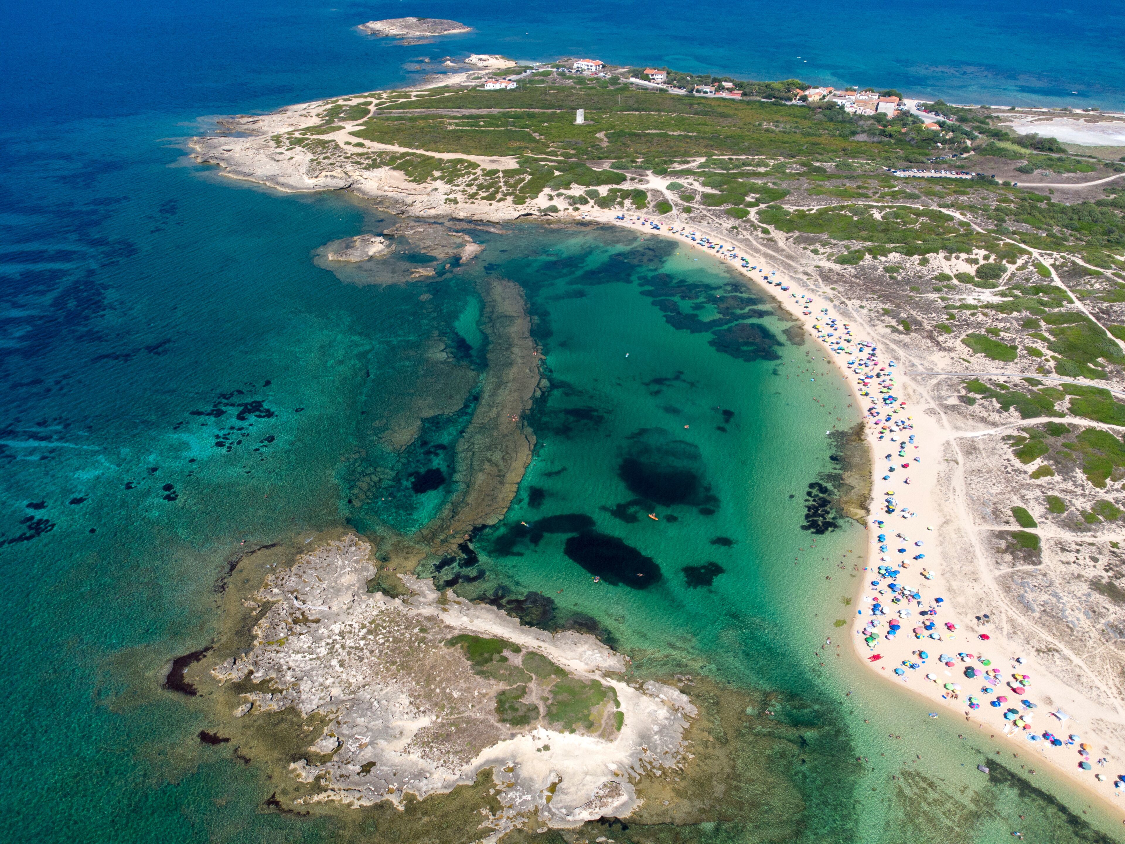 An aerial view of a coastal area with clear turquoise waters, a sandy beach, and scattered buildings.