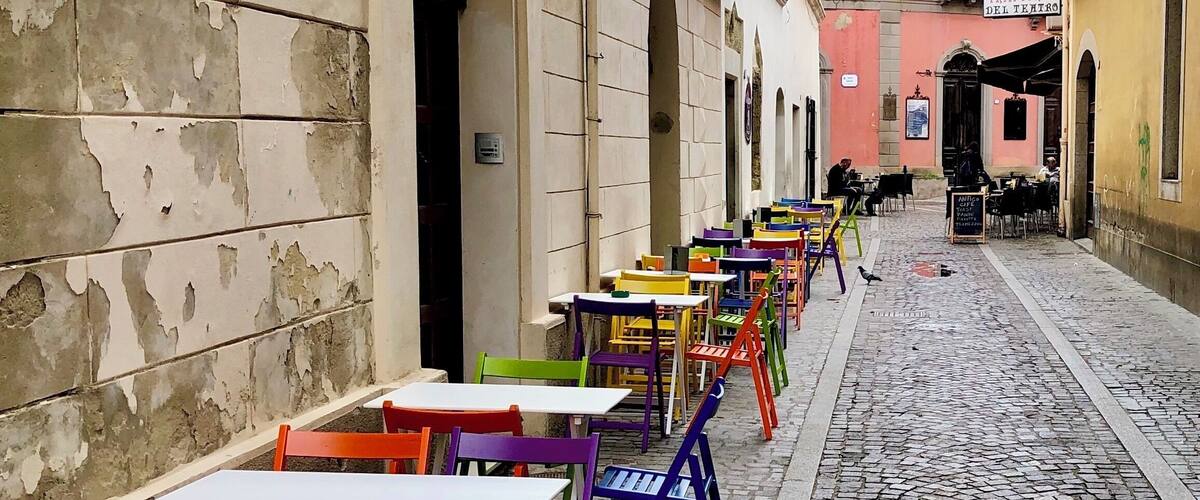 A narrow cobblestone street lined with colorful outdoor tables and chairs, suggesting a vibrant café or restaurant area.