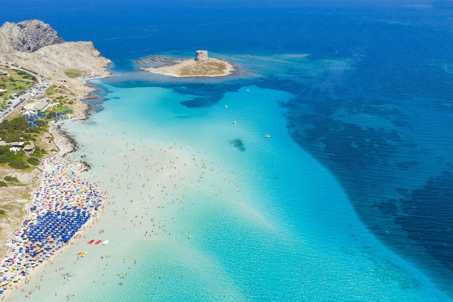 A coastal beach with clear turquoise waters, a rocky outcrop, and a small island with a tower.