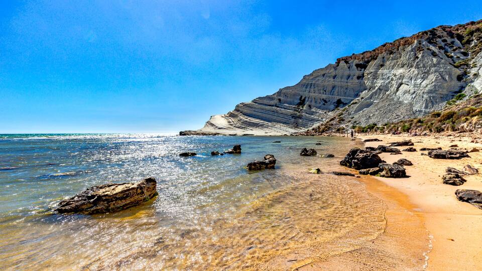 A beach with clear shallow waters, rocky outcrops, and a steep cliff.