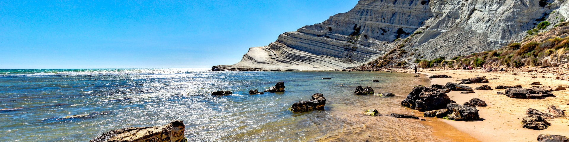 Uma praia com águas cristalinas e rasas, afloramentos rochosos e um penhasco íngreme.
