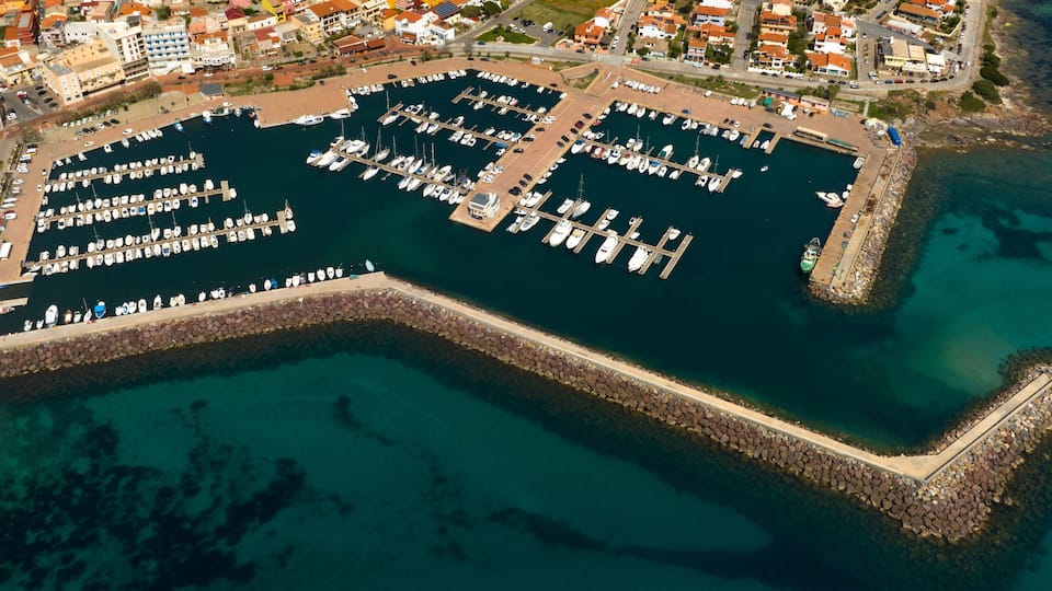 An aerial view of a marina with numerous boats docked, surrounded by residential buildings and a rocky breakwater.