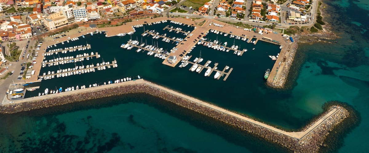 An aerial view of a marina with numerous boats docked, surrounded by residential buildings and a rocky breakwater.