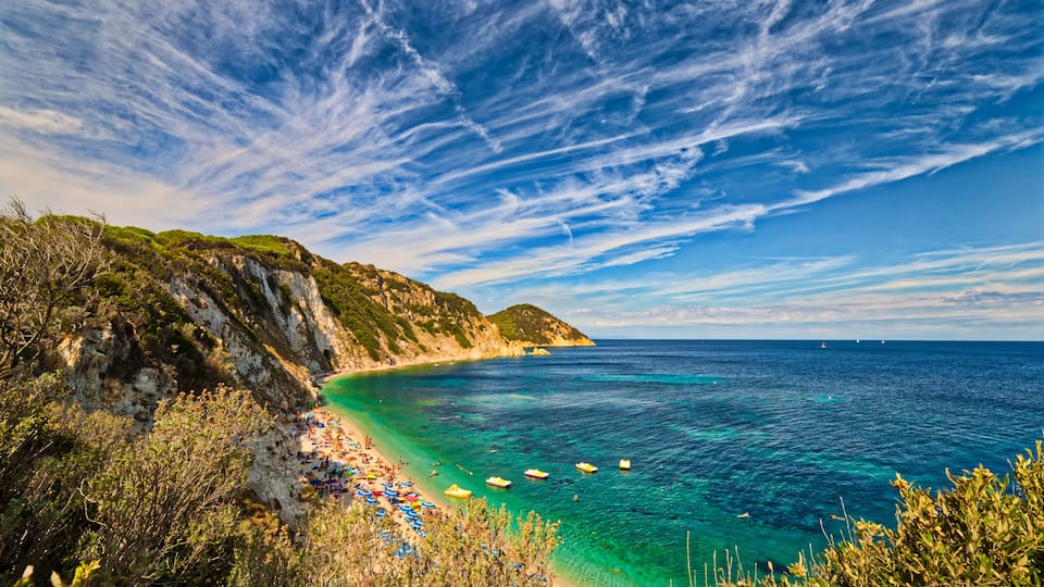 A coastal landscape with a clear blue sea, a sandy beach, and rocky cliffs.