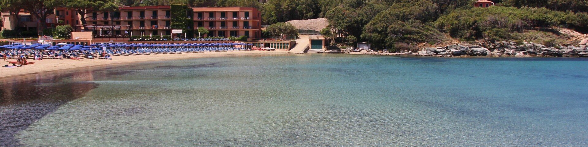 A coastal beach with clear water, a row of blue umbrellas, and a building with red roofs in the background.