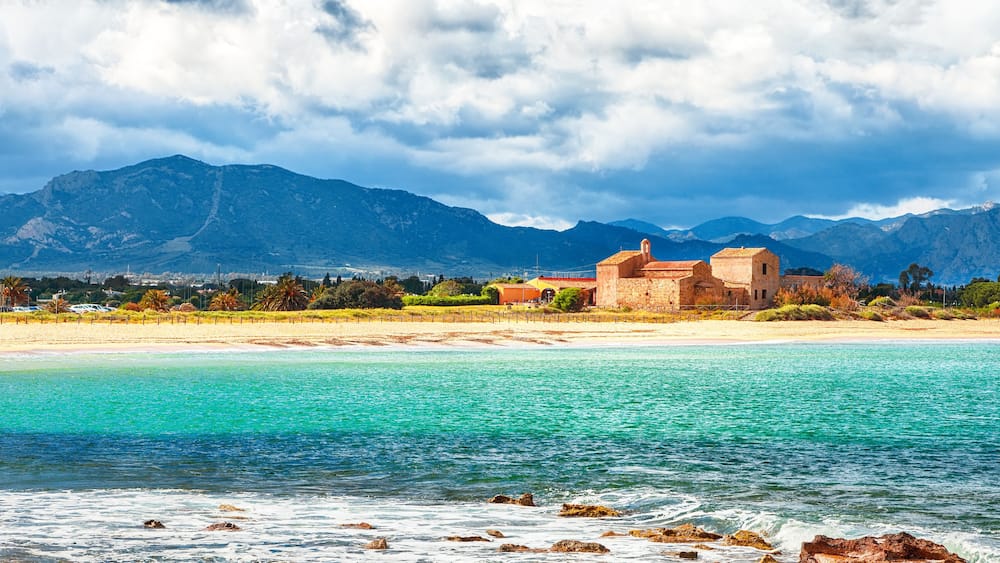 A coastal landscape with a beach, clear turquoise waters, and mountains in the background.