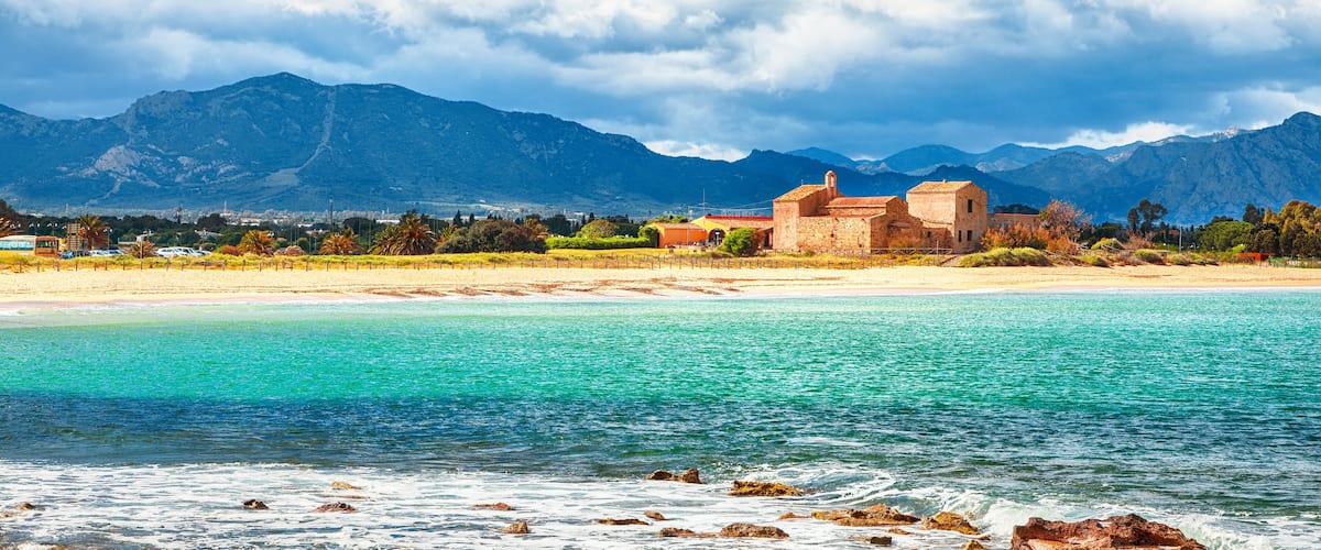 A coastal landscape with a beach, clear turquoise waters, and mountains in the background.