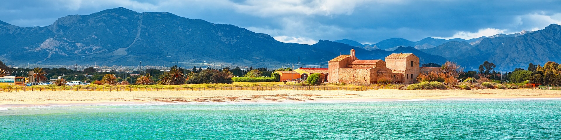 A coastal landscape with a beach, clear turquoise waters, and mountains in the background.
