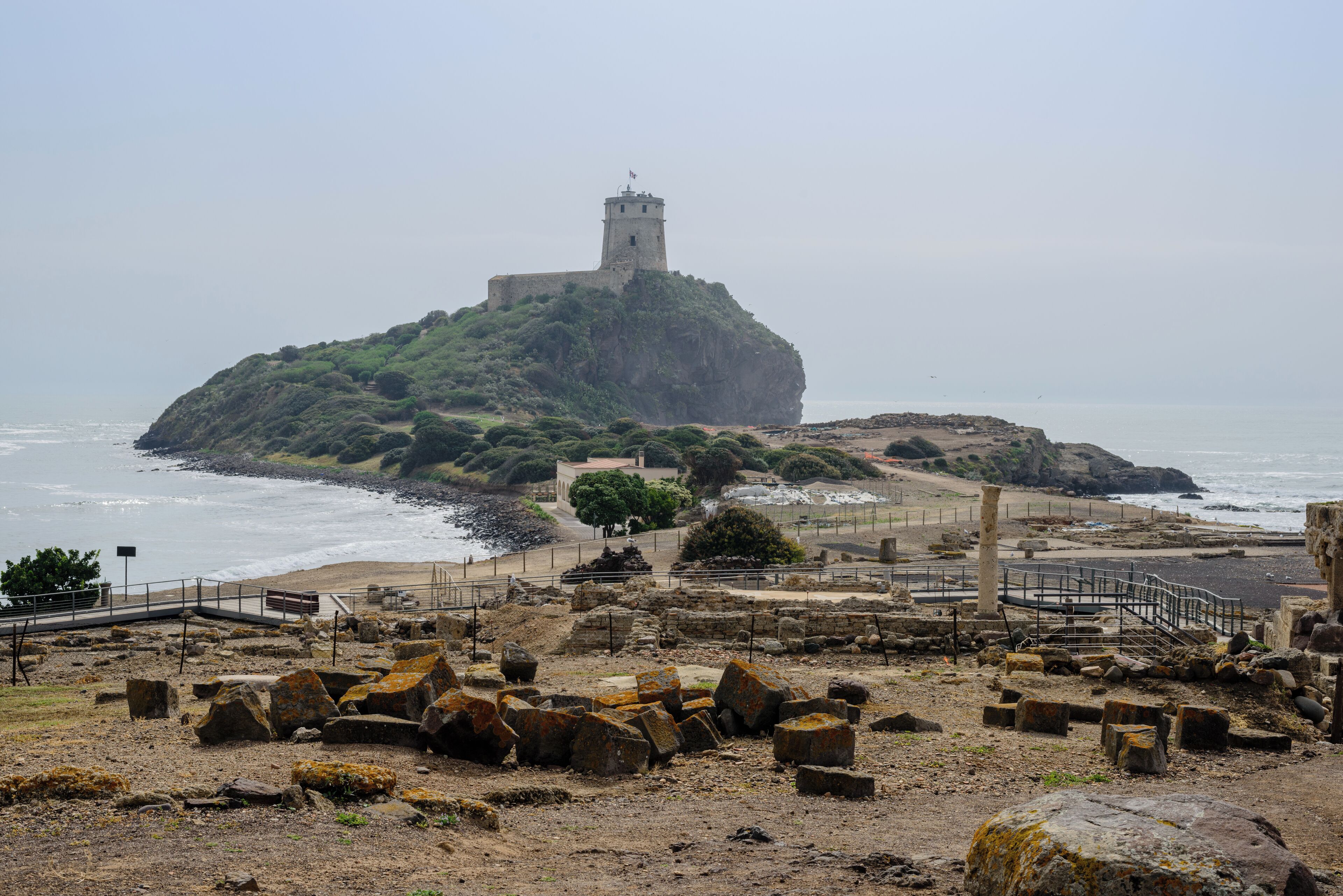 A coastal fortification with a tower on a rocky island, surrounded by ruins and a beach.