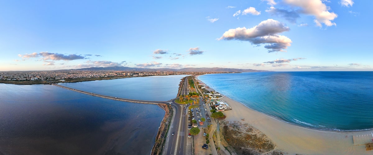 An aerial view of a coastal city with a prominent road leading to a beach, a bridge connecting two land masses, and a clear blue sky.