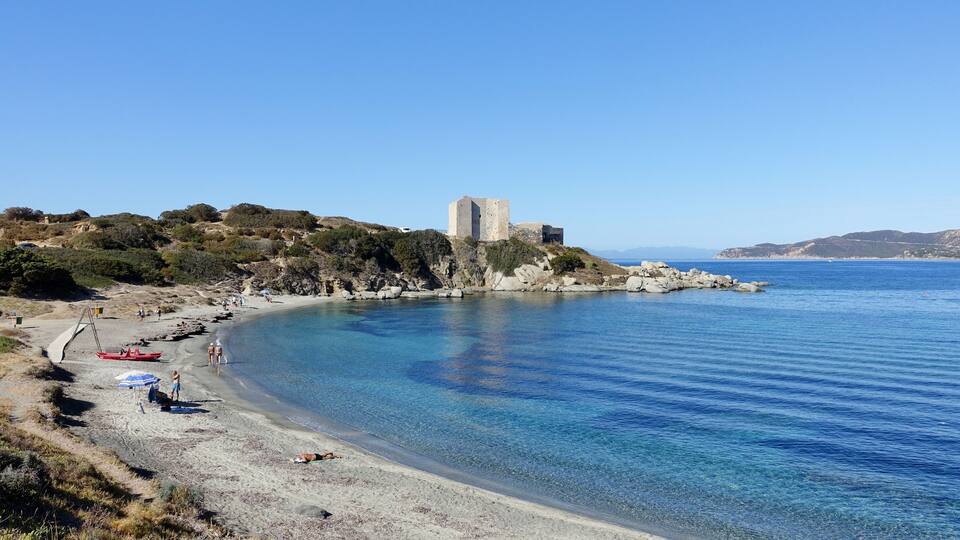 A coastal landscape with a clear blue sea, a sandy beach, rocky outcrops, and a historic stone tower on a hill.