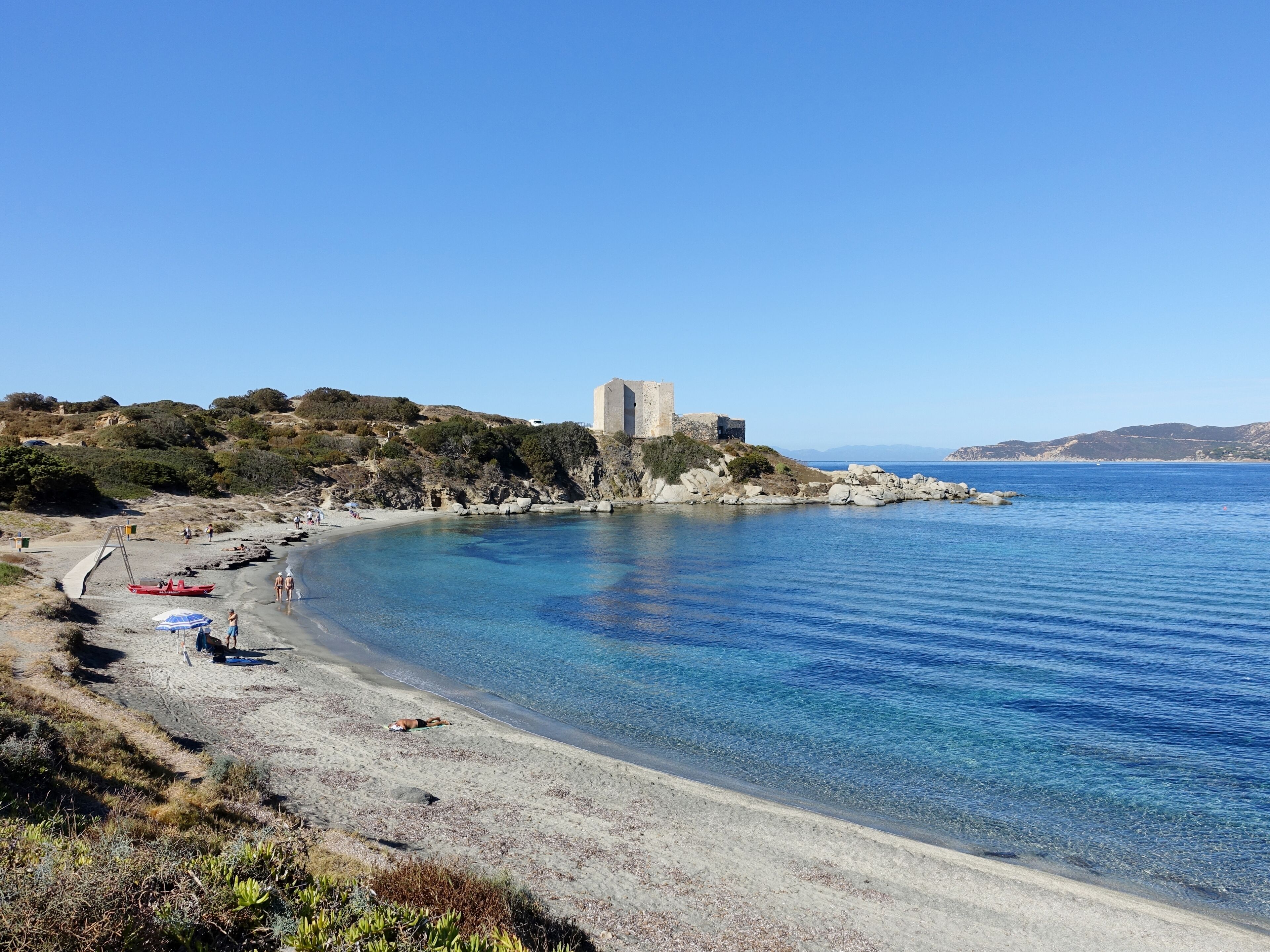 A coastal landscape with a clear blue sea, a sandy beach, rocky outcrops, and a historic stone tower on a hill.